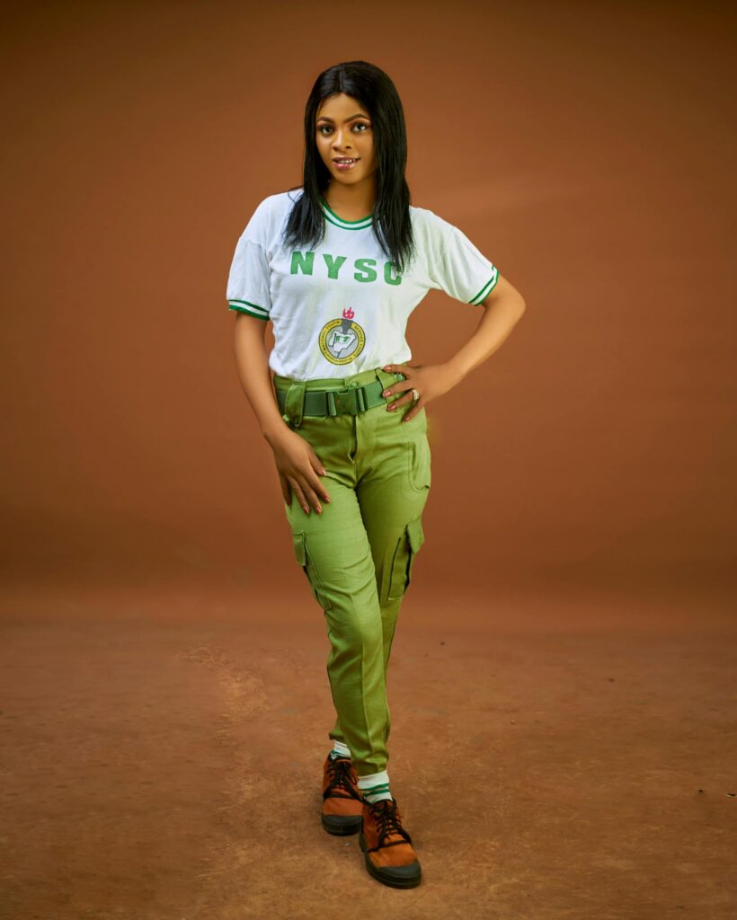 Young woman in NYSC uniform posing against brown backdrop.